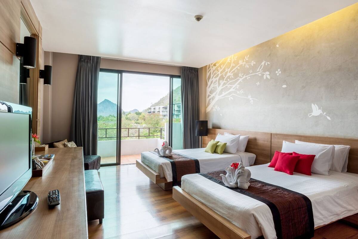 A hotel room with two beds, a TV, a desk, and a balcony with a view of greenery and buildings.