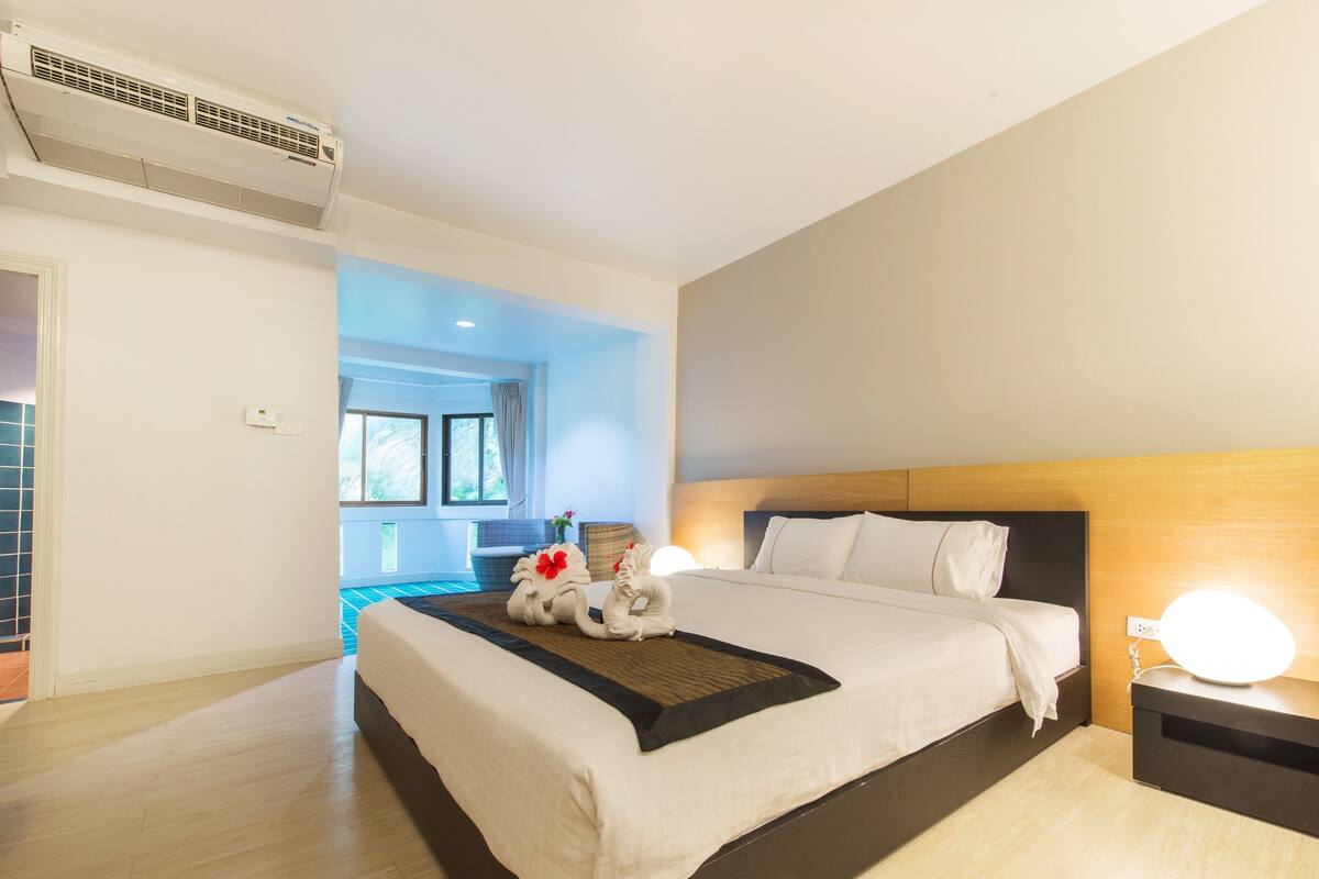 A modern hotel room with a large bed, a bedside table with a lamp, and a view of the outside through a window.