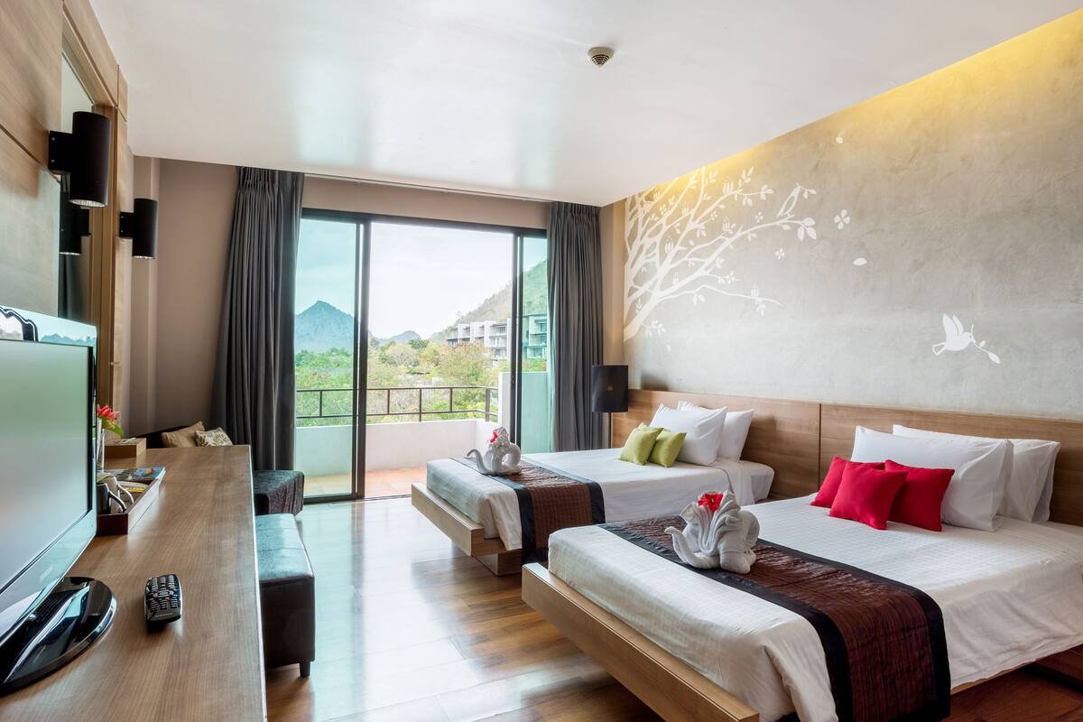 A hotel room with two beds, a desk with a TV, and a balcony with a view of the mountains.