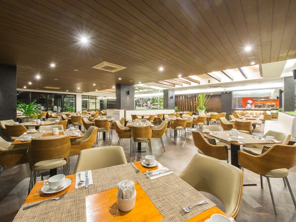 Un grand espace dédié à la salle à manger, avec plusieurs tables et chaises, une cuisine à l’arrière-plan et de grandes fenêtres qui laissent entrer la lumière naturelle.