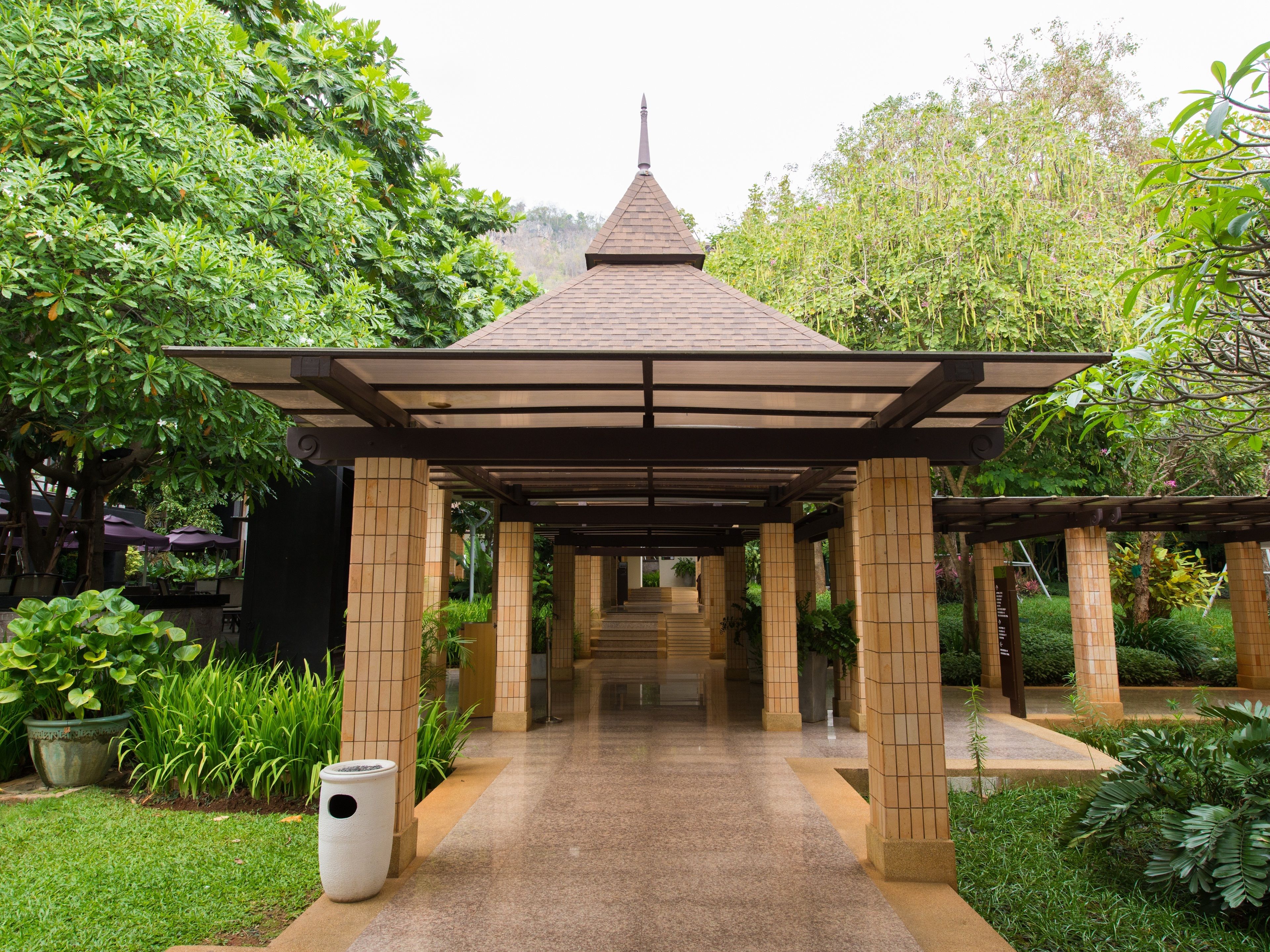 A covered pavilion with a tiled floor, surrounded by lush greenery and a tiled pathway leading to it.