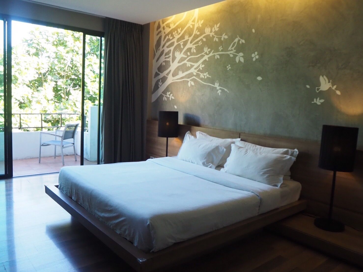 A modern hotel room with a large bed, a balcony with a chair, and a wall mural.