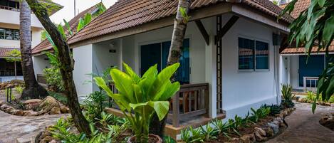 Une maison avec un toit en tuiles, entourée d’un jardin où poussent différentes espèces de plantes et bordée d’un chemin en pierre.