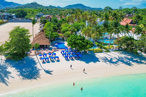 Vista aérea de un complejo turístico con piscina, sombrillas de playa y palmeras.