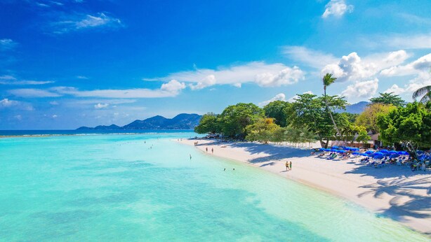 Une plage à l’eau turquoise et limpide, avec un rivage sablonneux et une rangée de parasols bleus.