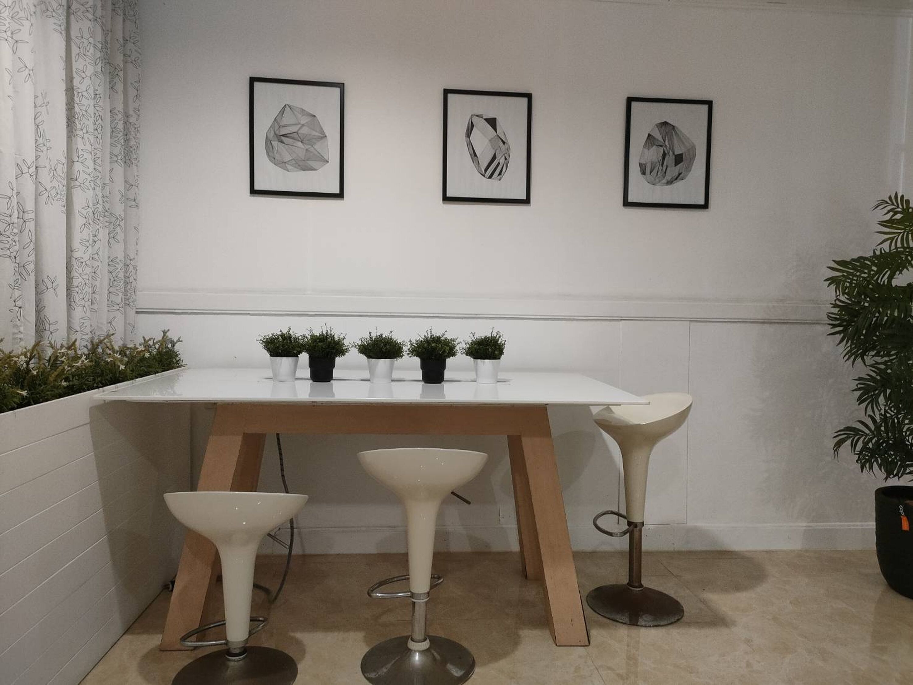 Une table blanche, ornée de quatre plantes en pot, de trois œuvres d’art encadrées et de trois tabourets de bar.