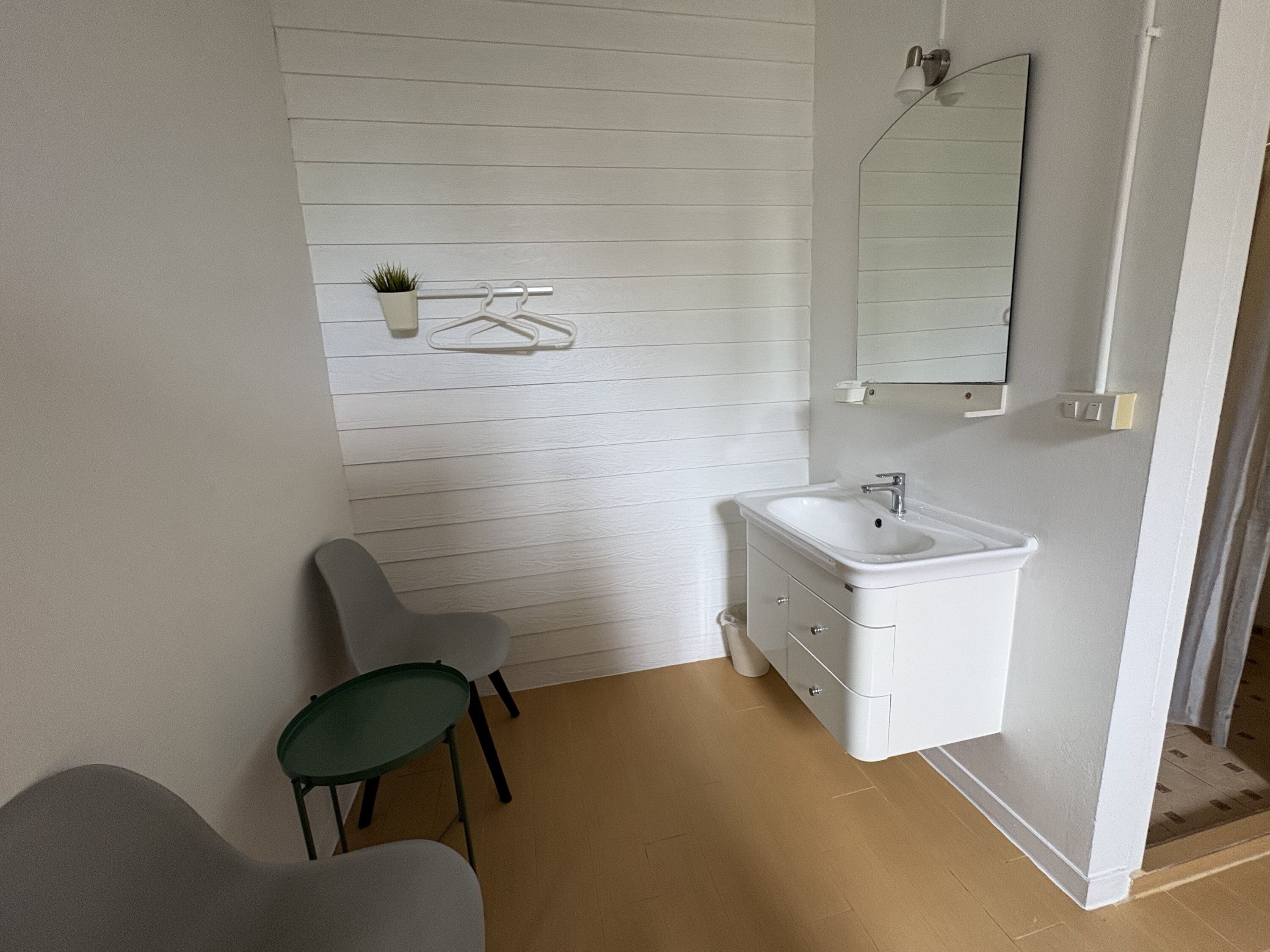 Ett badrum med ett vitt handfat, en spegel och en handdukshängare med en växt. Det finns två stolar och ett litet bord.