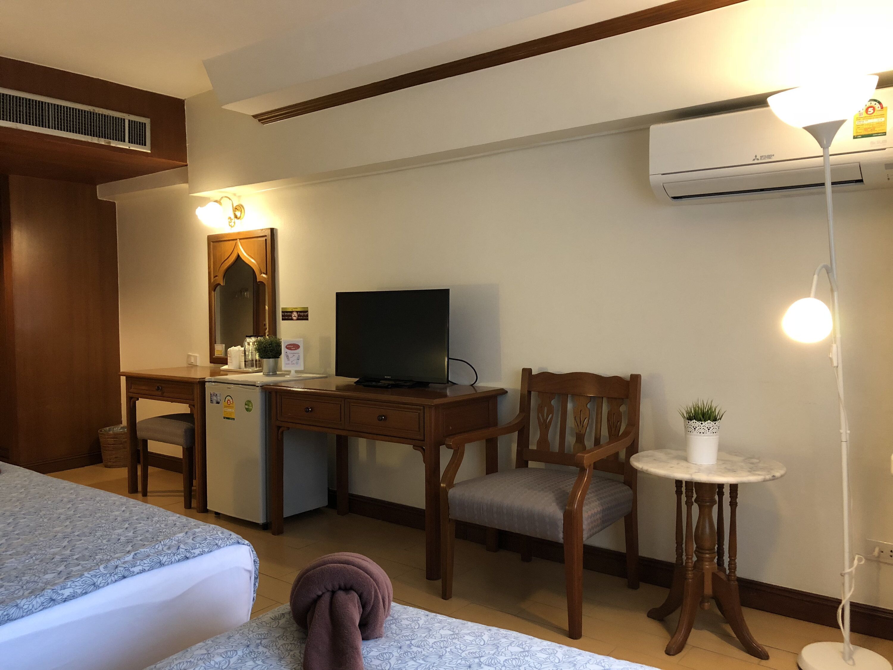 A hotel room with a bed, a desk with a television, a chair, a small table with a plant, and an air conditioning unit.