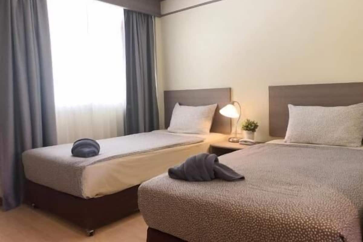 Habitación de hotel con dos camas, una mesita de noche con lámpara y una ventana con cortinas.