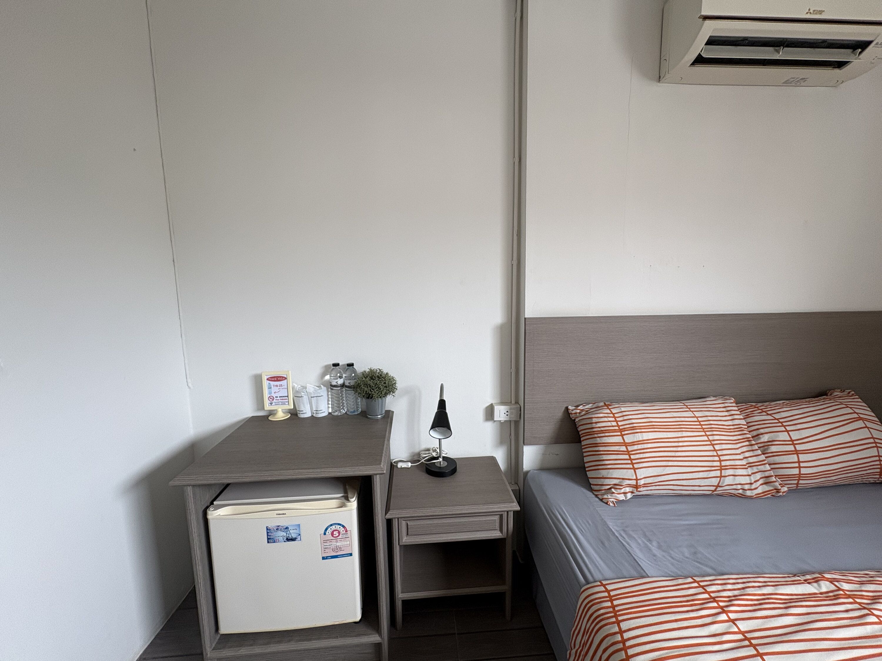 Ein Hotelzimmer mit einem Bett, einem kleinen Schreibtisch, einem Kühlschrank, einem Nachttisch und einer Wand-Klimaanlage.