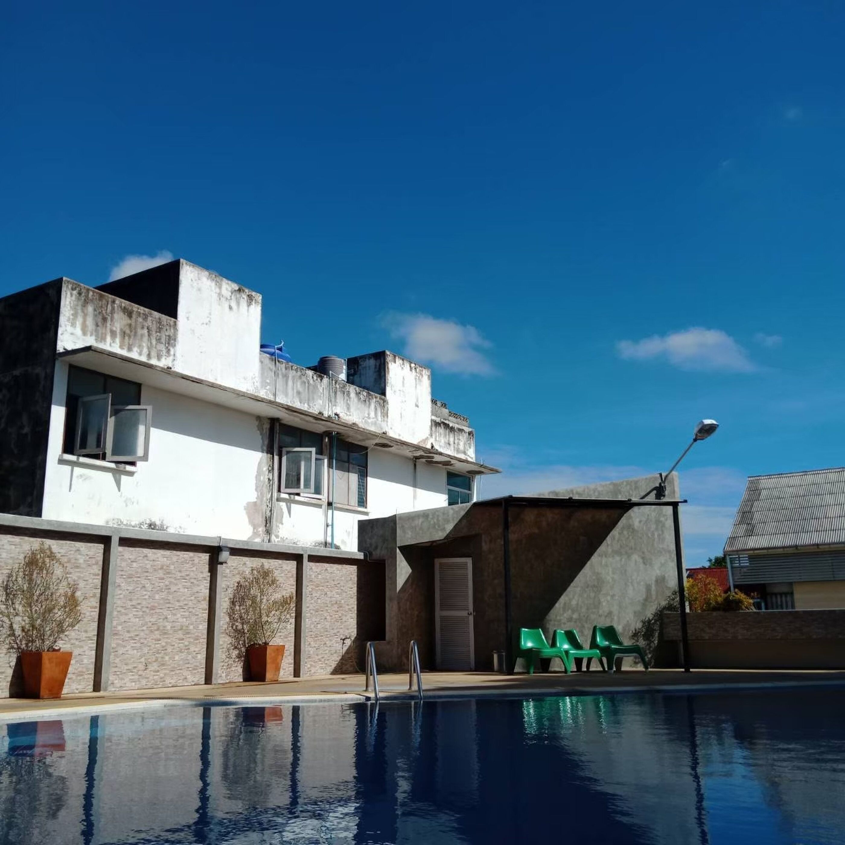 A swimming pool with a clear blue sky, a building with a white facade, and green chairs.