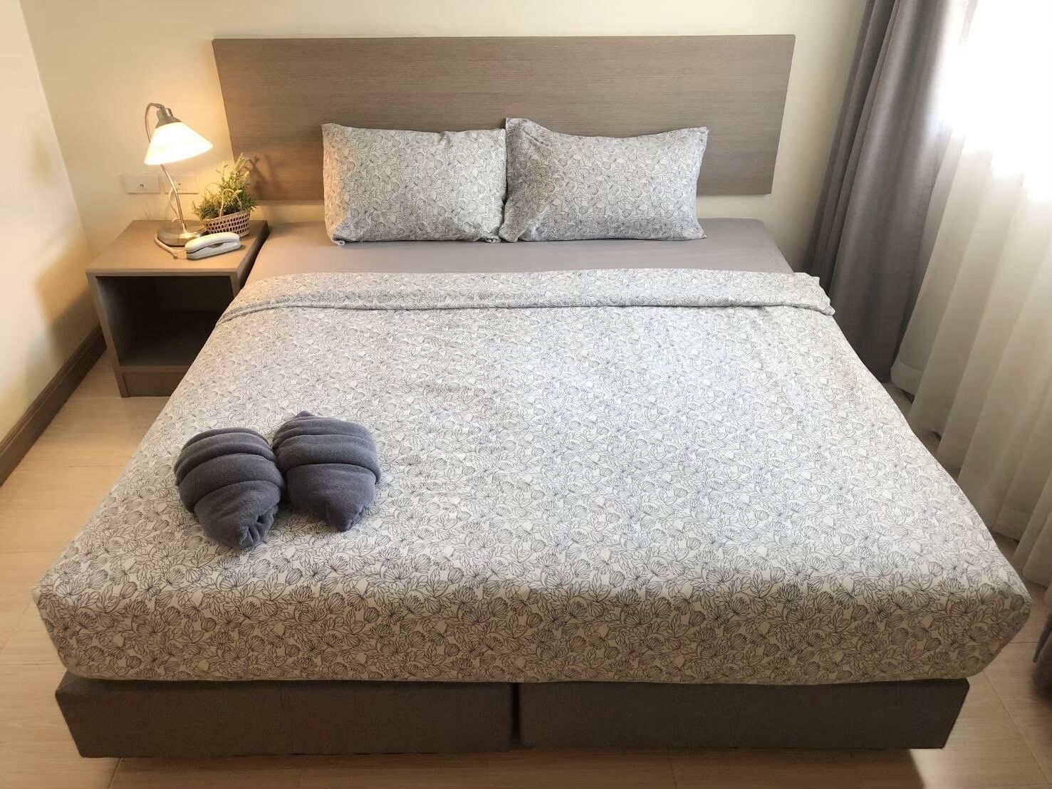 Un lit bien fait, recouvert d’une couvre-lit à motifs floraux, avec deux oreillers, une lampe de chevet et une petite plante posée sur une table de chevet.