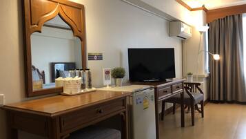 A hotel room with a wooden desk, a television, a refrigerator, a bench, a mirror, a wall-mounted air conditioner, a potted plant, and a window with curtains.