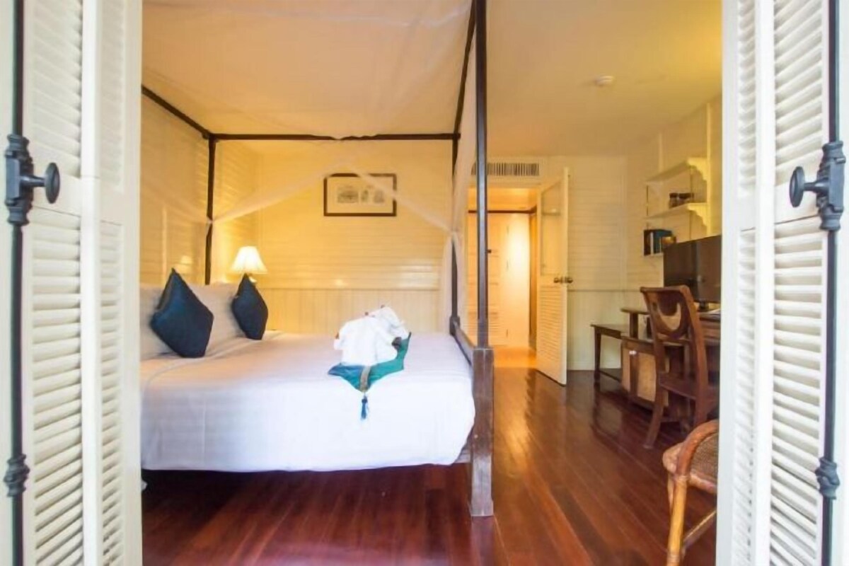 Un dormitorio con cama, almohadas, una mesita de noche y una lámpara. El suelo es de madera y hay una puerta que da a otra habitación.