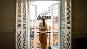 A person standing at a glass door with white shutters, looking outside. - Buddy Lodge (Bangkok)