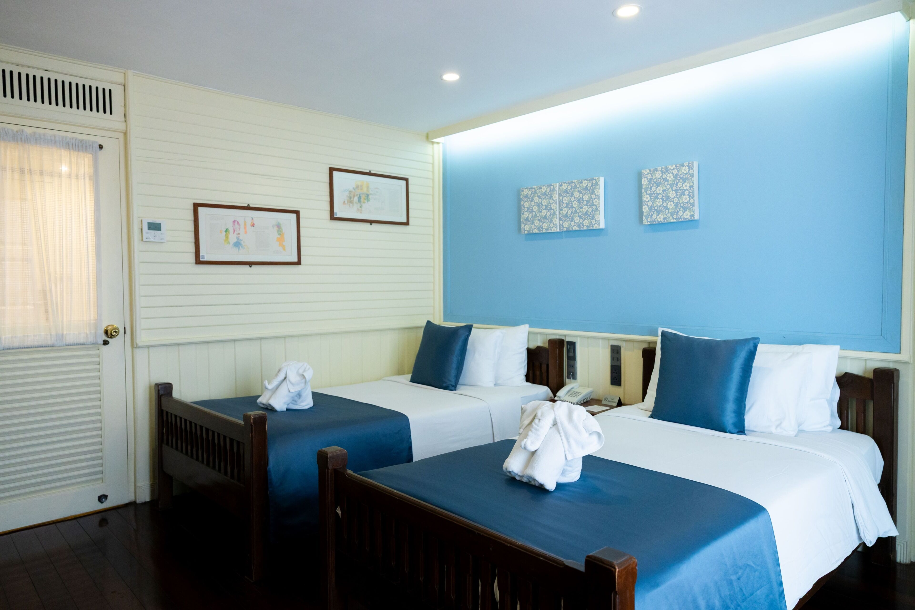 A hotel room with two beds, a wooden chair, a door with a window, and wall-mounted artwork.