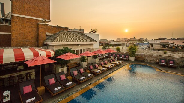 A rooftop pool area with lounge chairs and umbrellas, a brick building, and a sunset in the background. - Buddy Lodge (Bangkok)