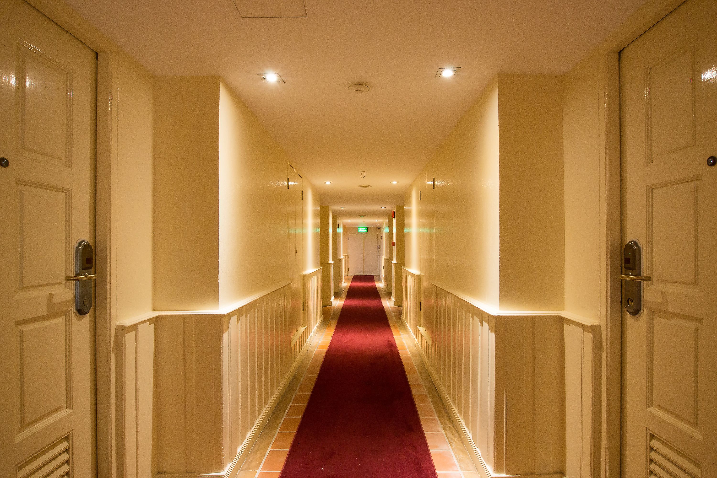 Un pasillo de hotel con alfombra roja, paredes color crema y puertas a ambos lados.