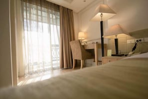 Relax in this stylish room with a large bed, desk, and lamp, offering a comfortable workspace and a large window with sheer curtains.
