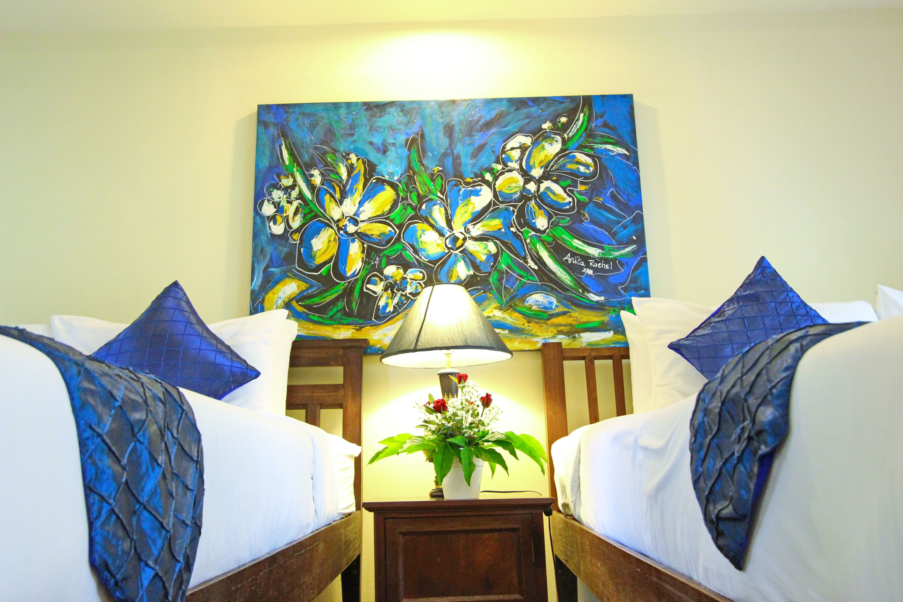 A hotel room with two beds, each with white linens and blue pillows. A painting of flowers hangs above the beds, and a nightstand with a lamp and a vase of flowers is in the center.
