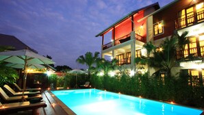 A swimming pool with lounge chairs and umbrellas, surrounded by lush greenery and a multi-story building with balconies. - Shewe Wana Suite Resort (Chiang Mai)