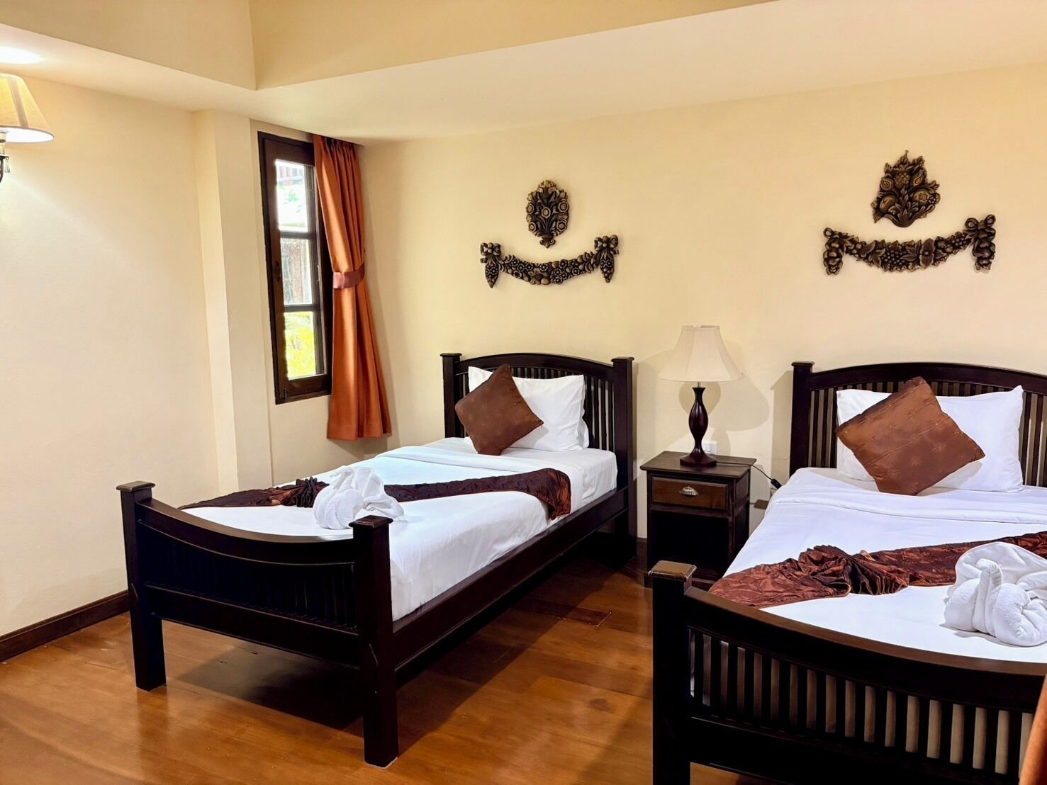 Habitación de hotel con dos camas de madera, una ventana con cortinas, una mesita de noche con lámpara y decoración en la pared.