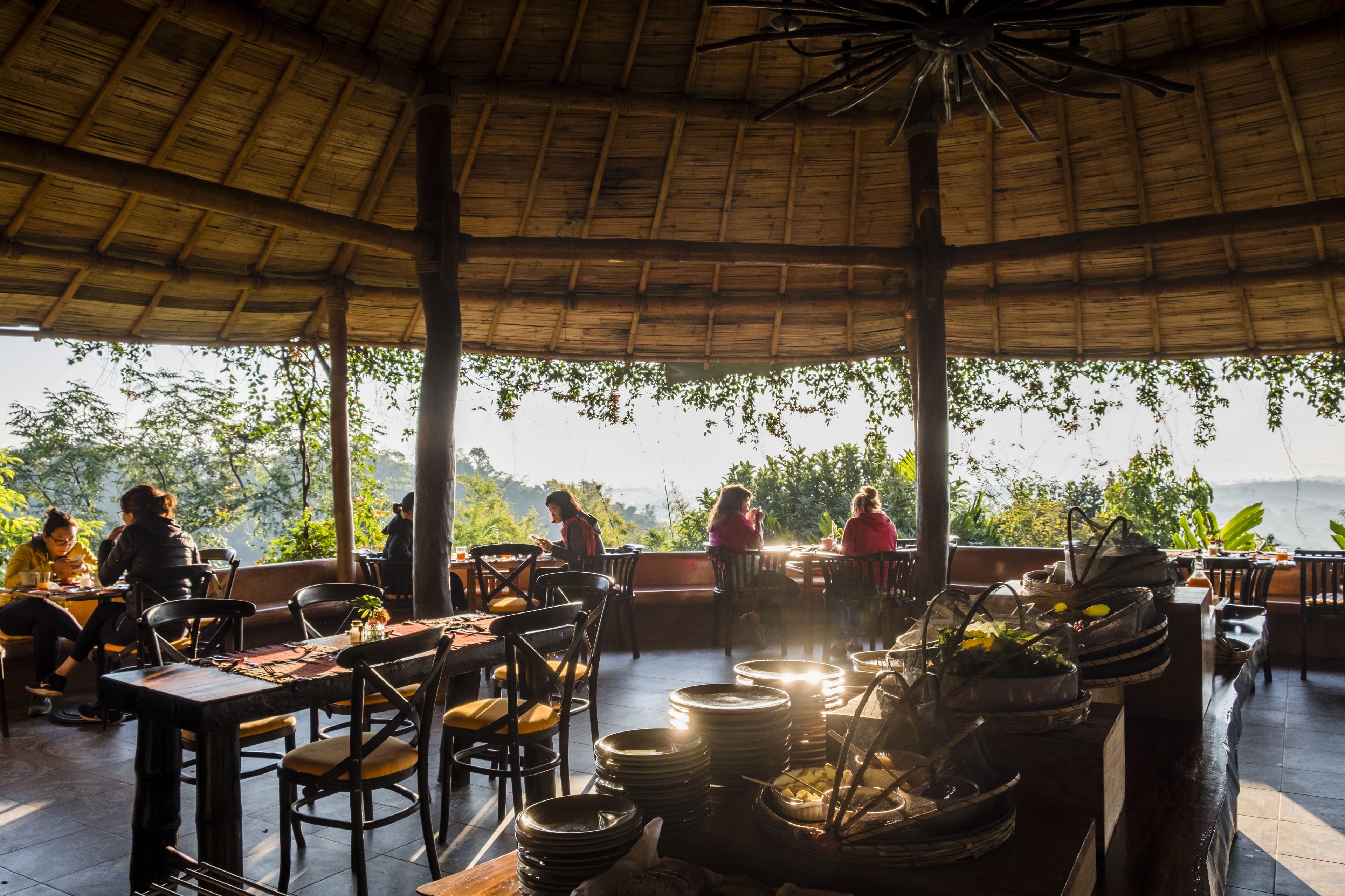 An outdoor dining area with tables and chairs, surrounded by a thatched roof structure.