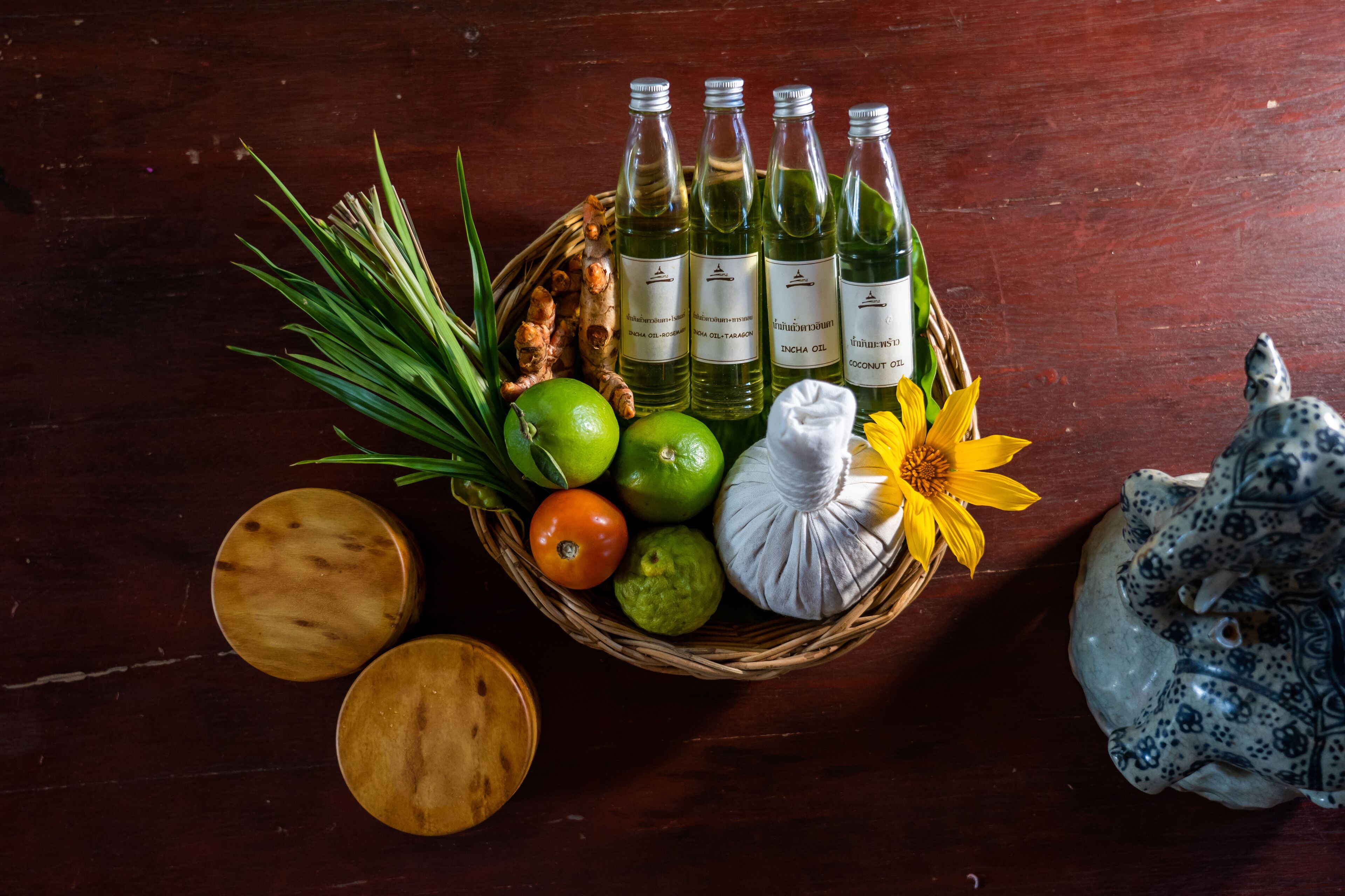 Une table en bois sur laquelle se trouve un panier contenant des bouteilles d’huile, un citron, une tomate, une tête d’ail et une fleur jaune.