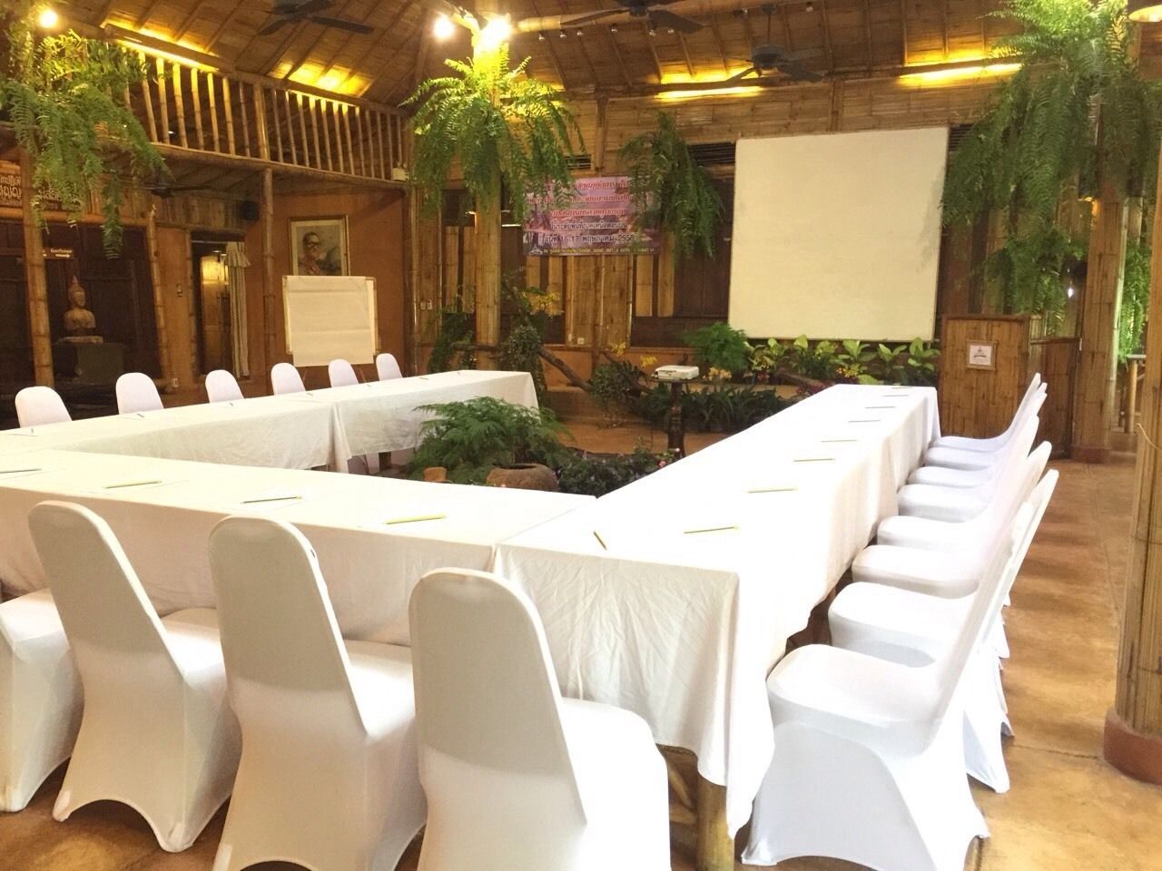 A conference room with long tables and white chairs, surrounded by wooden walls and indoor plants.