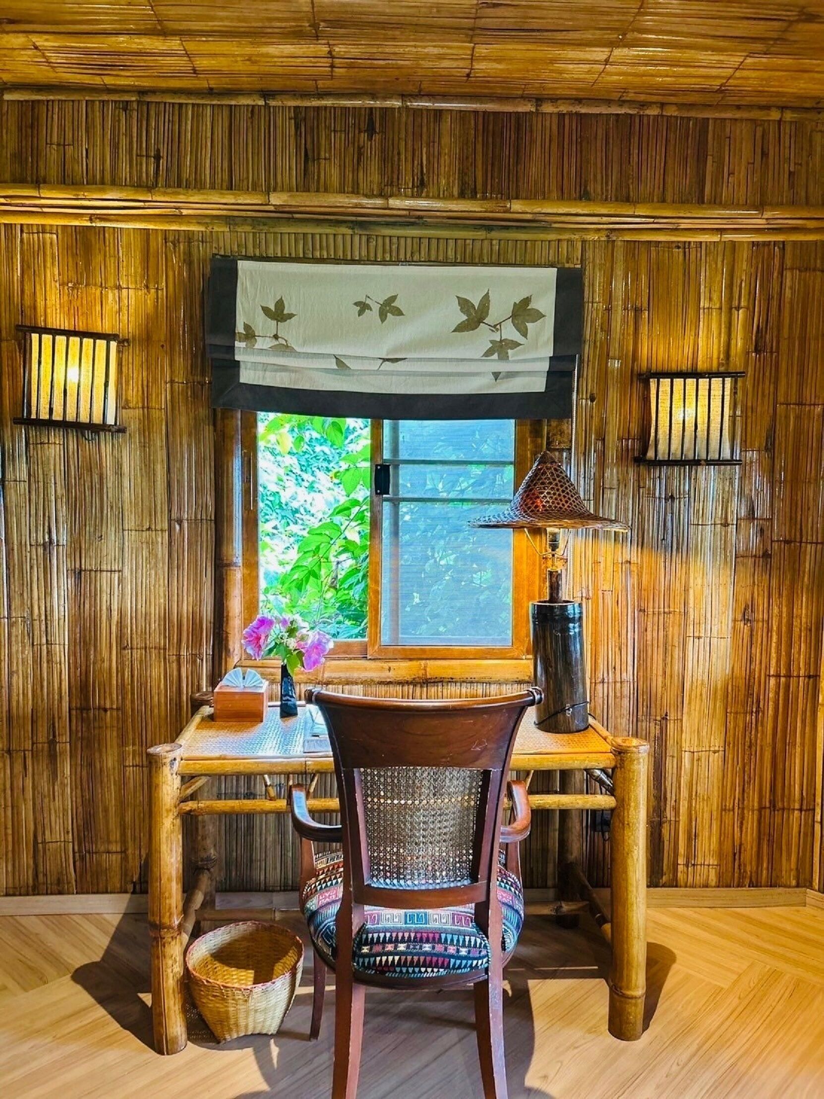 A bamboo-paneled room with a wooden desk, a chair, a window with a view of greenery, and a decorative hanging above the desk.