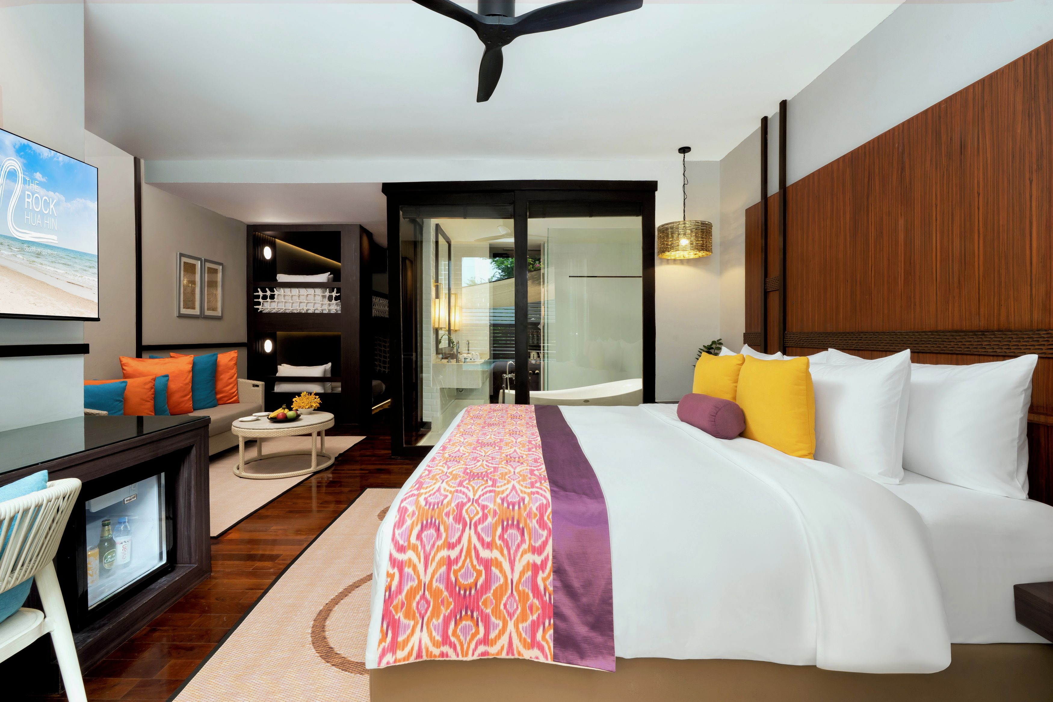 Quarto de hotel moderno com cama grande, escrivaninha, televisão e casa de banho com paredes de vidro.