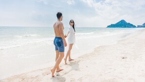Una pareja caminando de la mano por una playa arenosa con cielo despejado y una montaña distante.