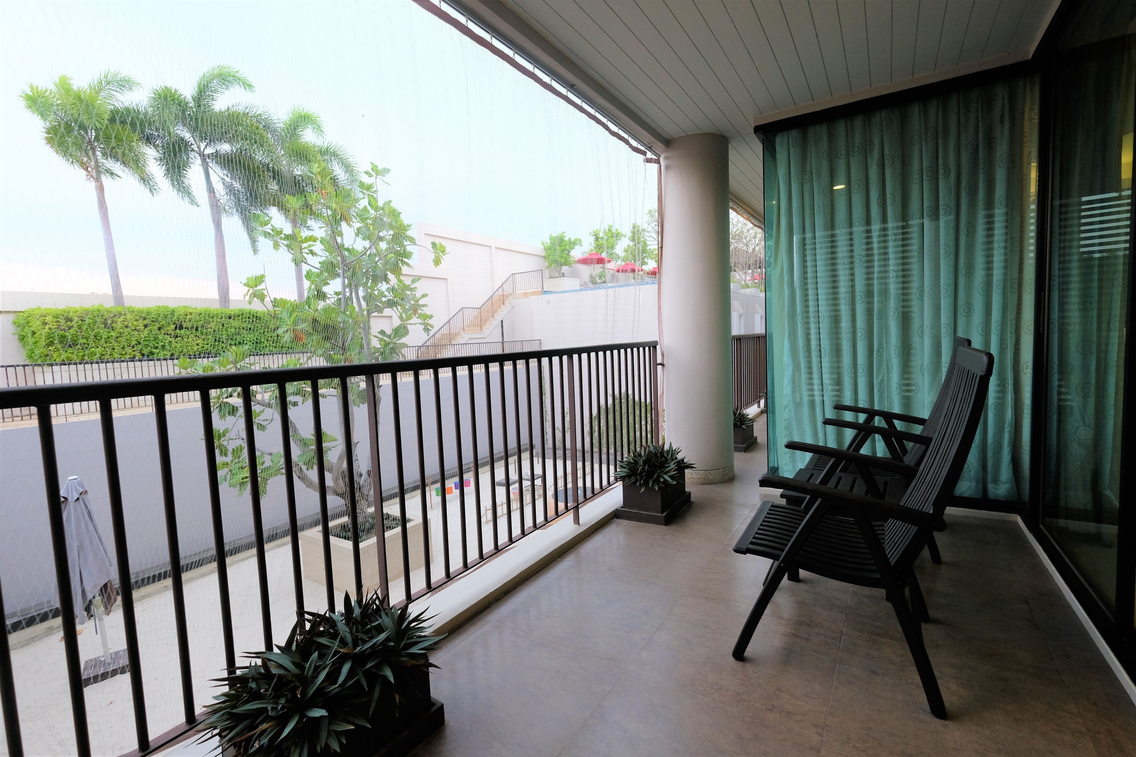 A balcony with a view of palm trees and a building. There are two chairs and a potted plant on the balcony.