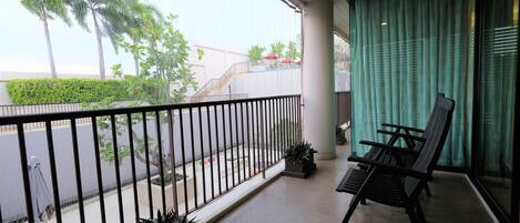 A balcony with a view of palm trees and a building. There are two chairs and a potted plant on the balcony.