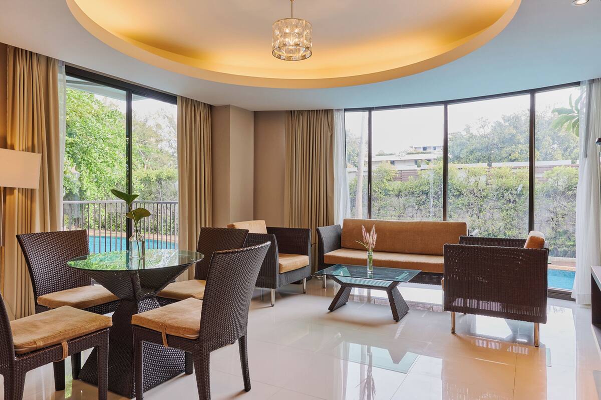 Quarto de hotel moderno com mesa de jantar de vidro, móveis de vime, sofá e grandes janelas com vista para áreas verdes e uma piscina.
