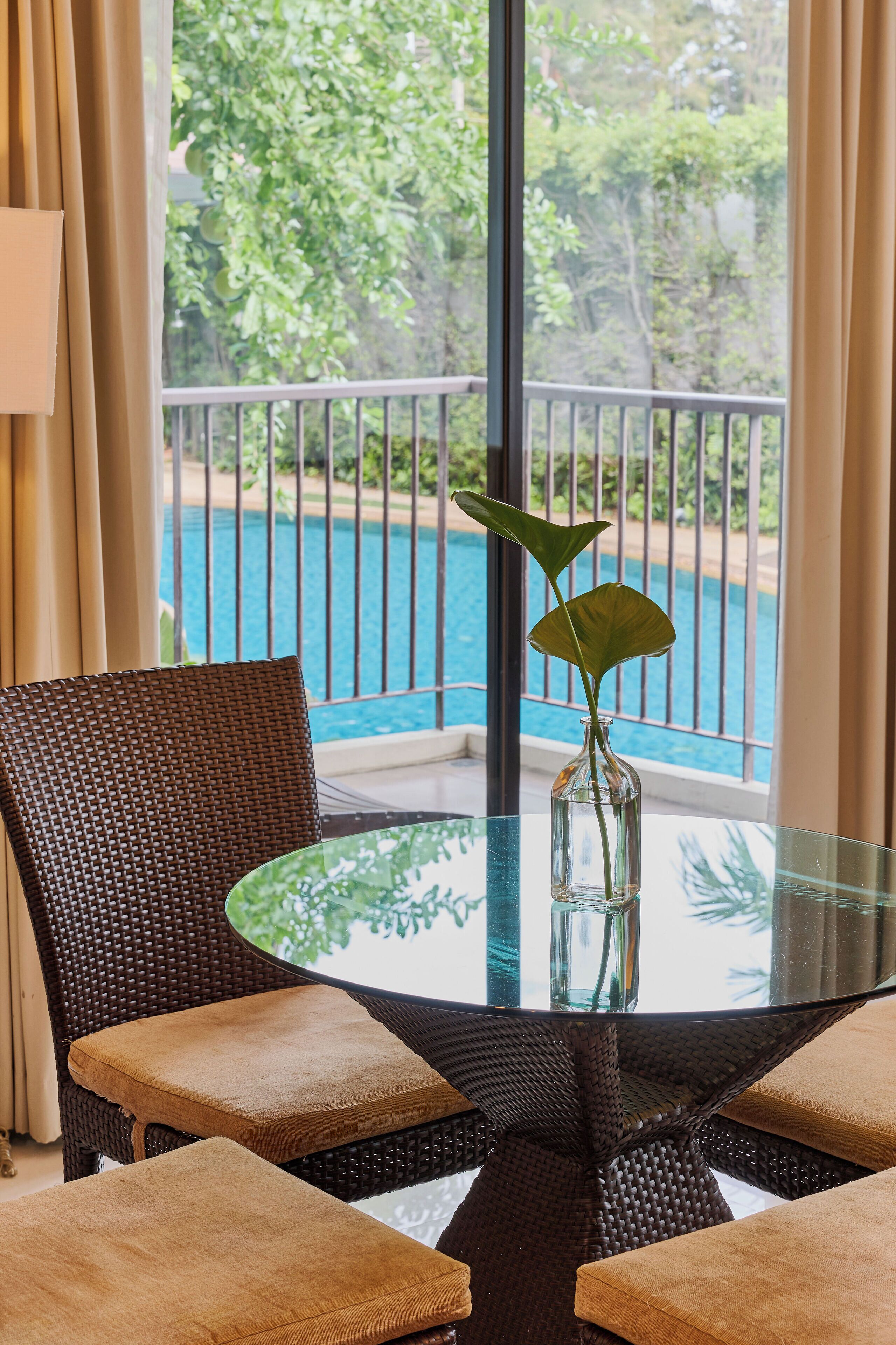 A glass table with a wicker chair and a cushion, a vase with a plant, and a view of a pool through a balcony with a railing.