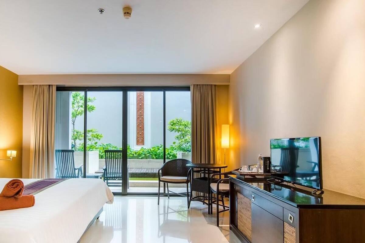 Quarto de hotel com cama, escrivaninha, televisão e varanda com plantas.