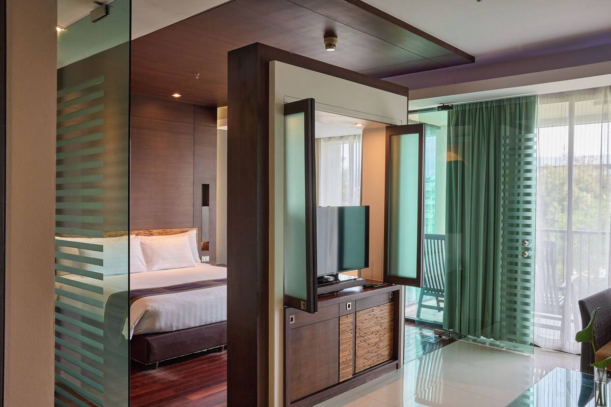Quarto de hotel moderno com uma cama grande, uma mesa de centro de vidro, uma TV sobre um móvel de madeira e vista para o exterior por uma porta de correr de vidro.