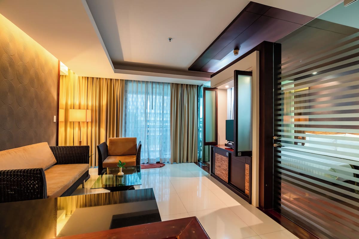 Quarto de hotel moderno com sofá, poltrona e mesa de centro de vidro. Há uma televisão em um armário de madeira, e uma porta de vidro deslizante com persianas.
