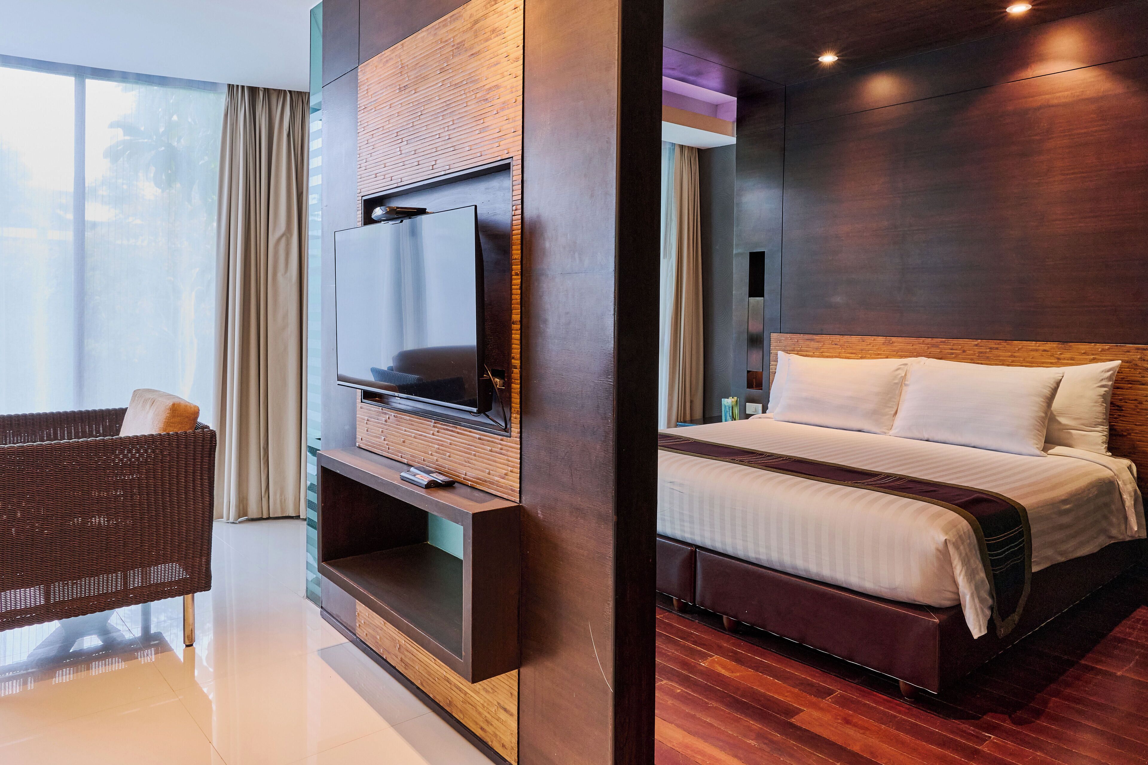 Une chambre d’hôtel moderne avec un grand lit, un téléviseur fixé sur un panneau en bois et un fauteuil en osier.