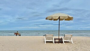A beach with two white lounge chairs and a beige umbrella, a horse and rider in the background, and the ocean. - Tara Mantra Cha Am Resort (Cha-am)
