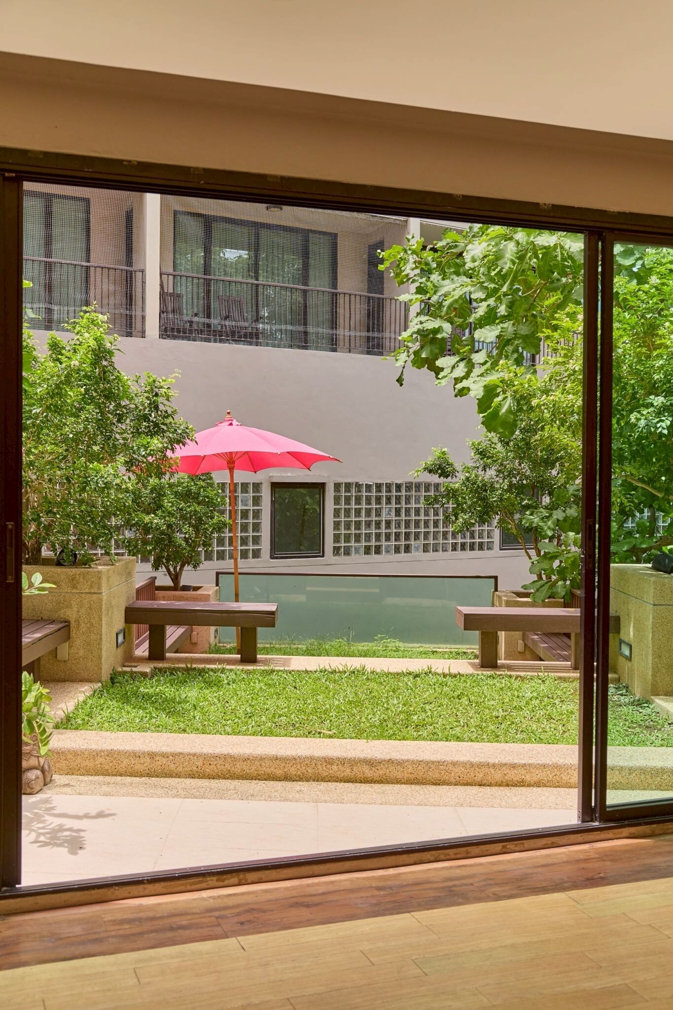 A balcony with a red umbrella, a small garden, and a view of a building with large windows.