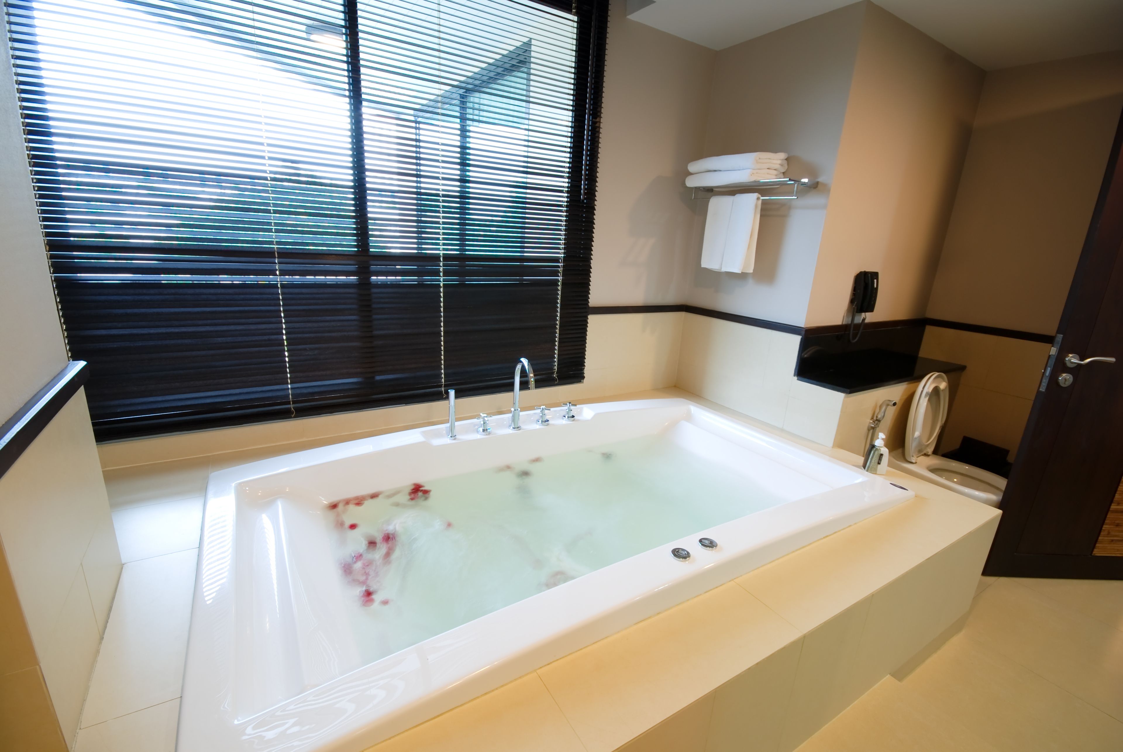 Un baño moderno con una bañera grande, un inodoro y un lavabo. Hay una ventana con persianas, un perchero y un teléfono en la pared.