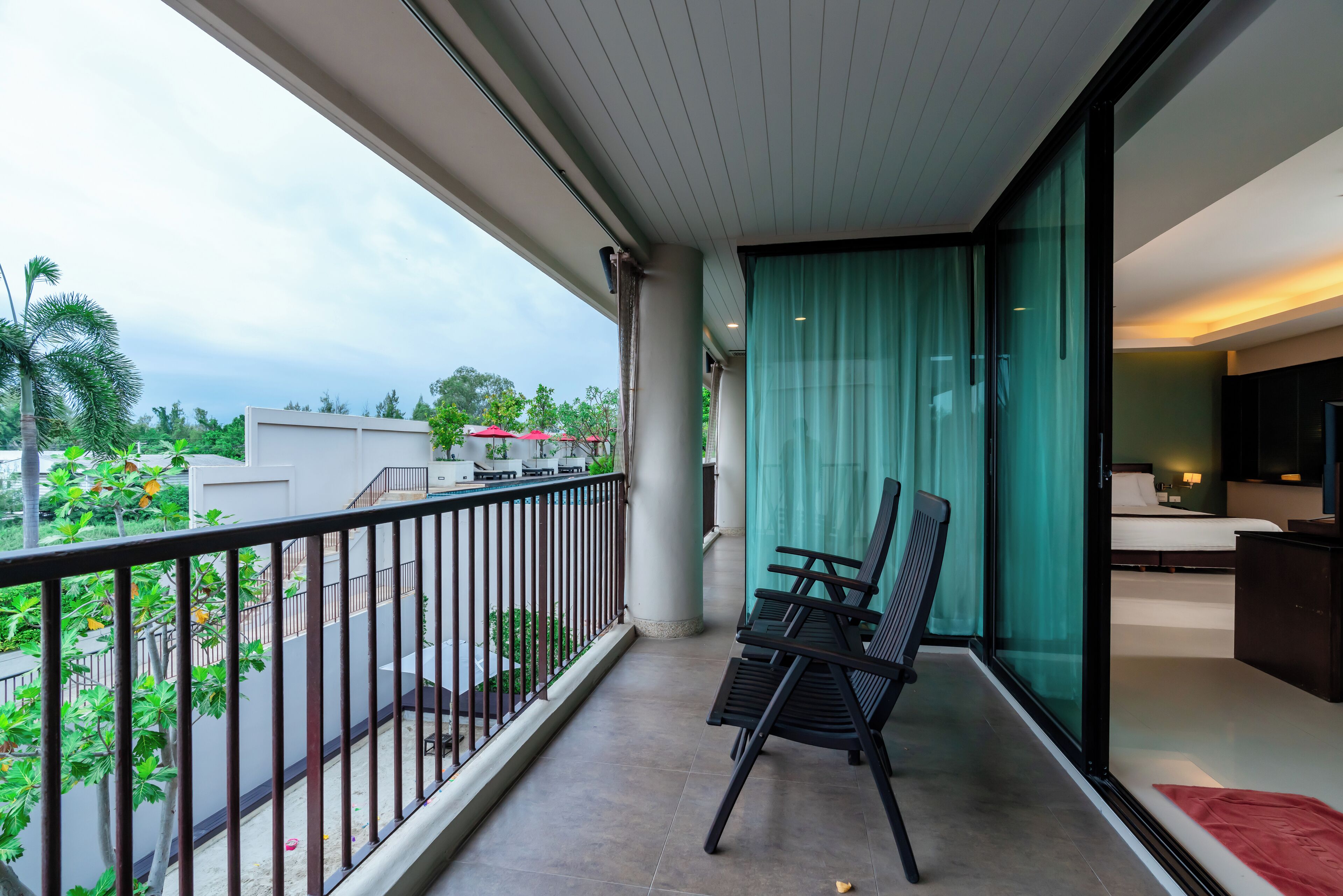 Un balcon avec vue sur la verdure et les bâtiments, un coin salon avec des chaises, et une chambre avec un lit et une télévision.