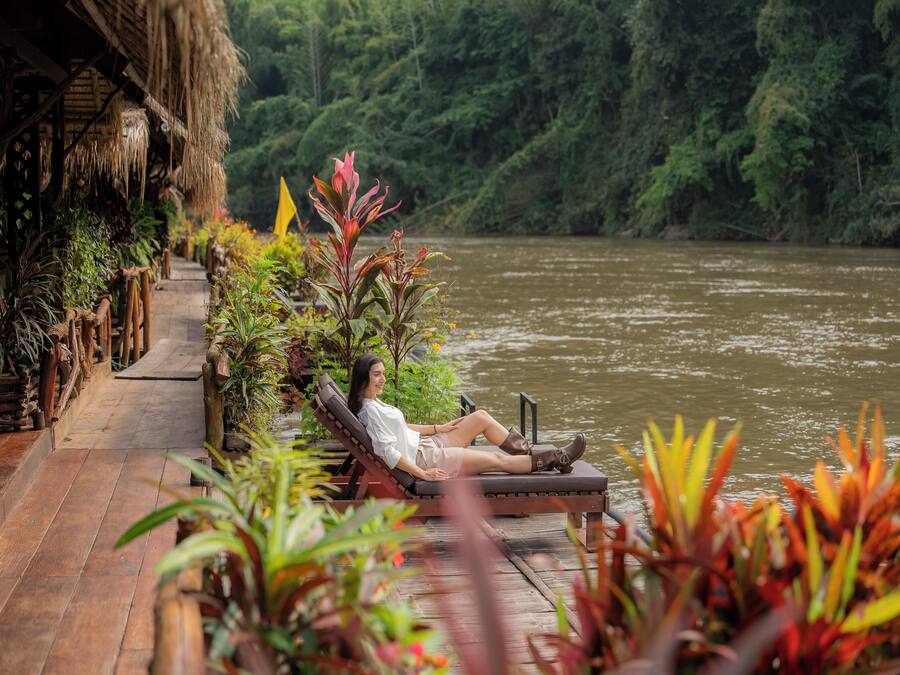 Une personne se détend sur une terrasse en bois, avec vue sur une rivière, entourée d’une végétation luxuriante et de plantes tropicales.