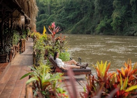 Una persona descansando en una terraza de madera con vista a un río, rodeada de exuberante vegetación y plantas tropicales.