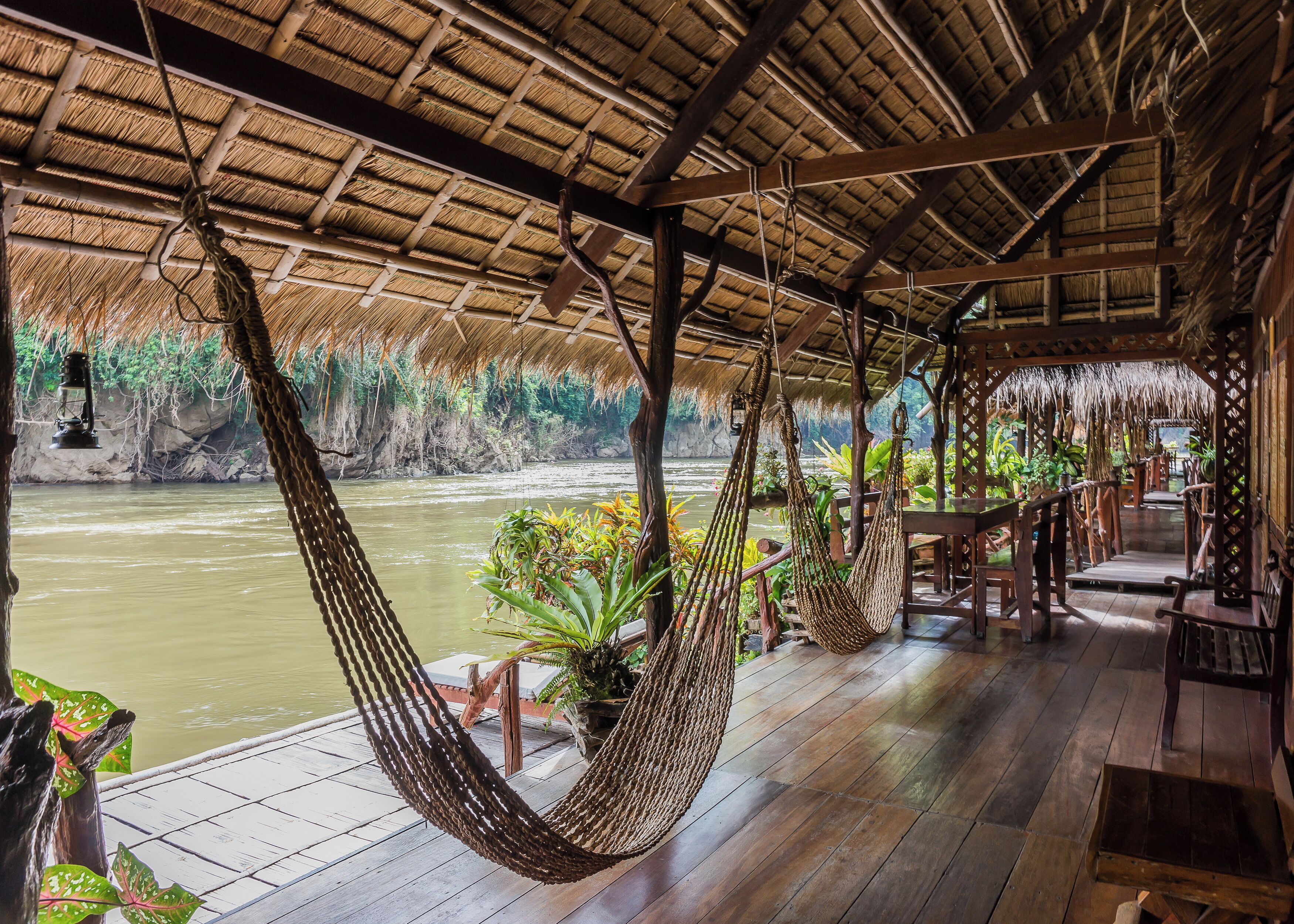 A wooden deck with a thatched roof overlooking a river, featuring hanging hammocks and potted plants.