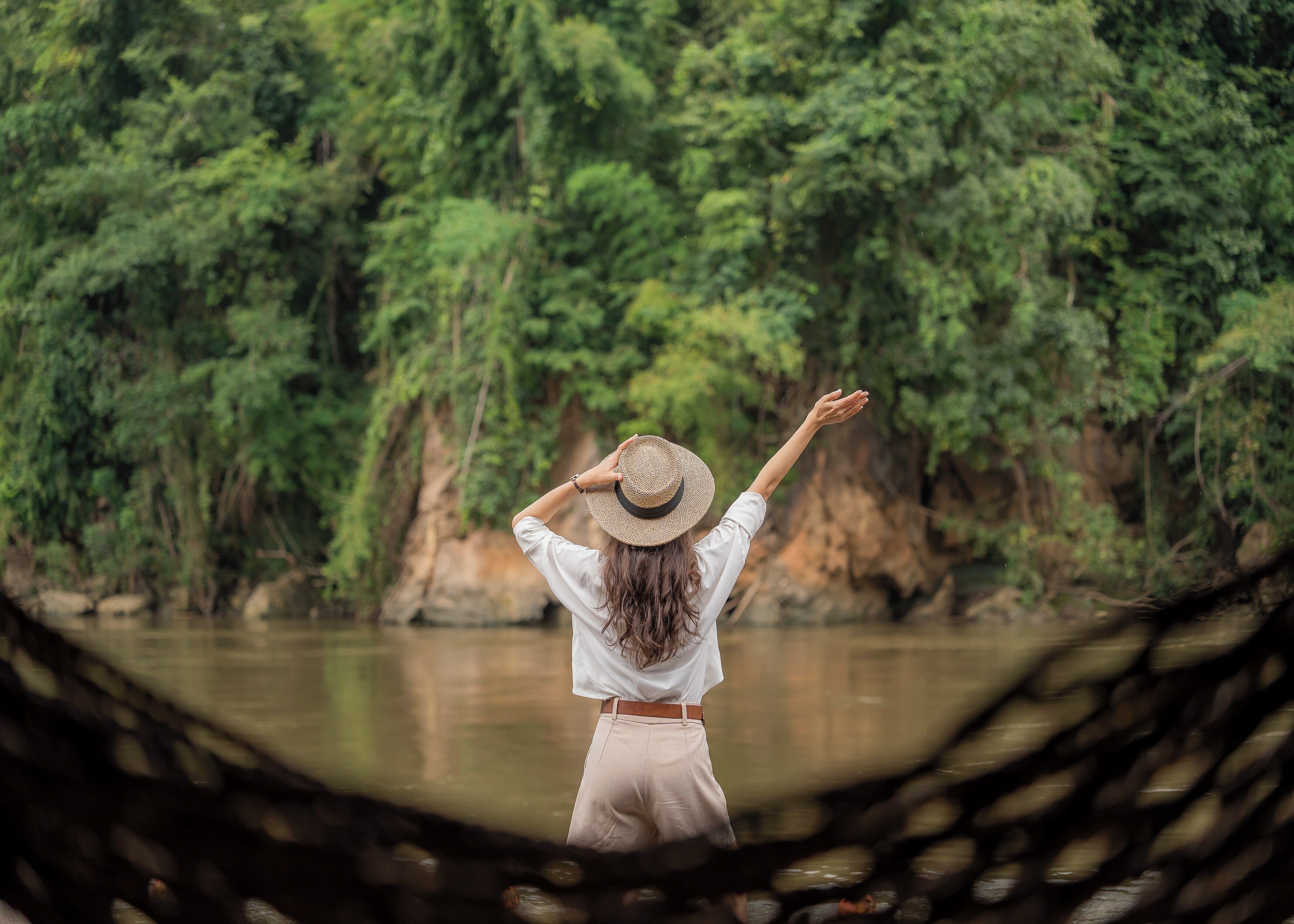 A person standing by a river, wearing a hat and a white shirt, with a natural landscape in the background.