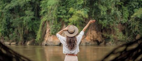 A person standing by a river, wearing a hat and a white shirt, with a natural landscape in the background.