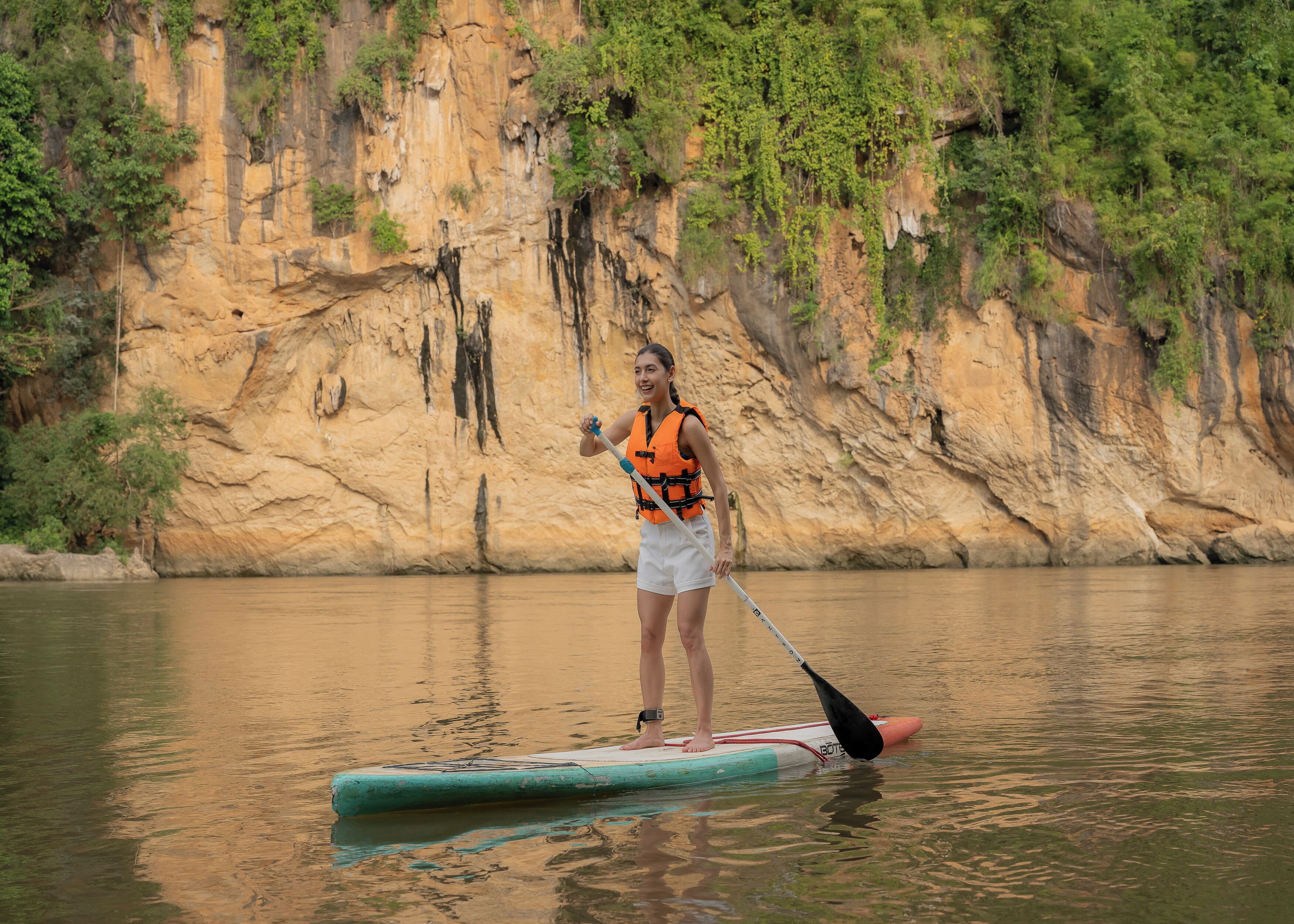 A person paddleboarding on a calm river with a rocky cliff in the background.